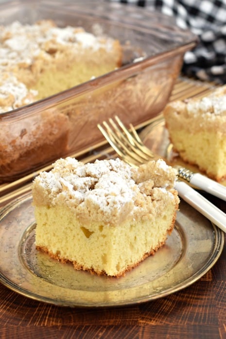 Two thick slices of crumb cake with powdered sugar on silver plates with white forks. Pan of Crumb cake in background.