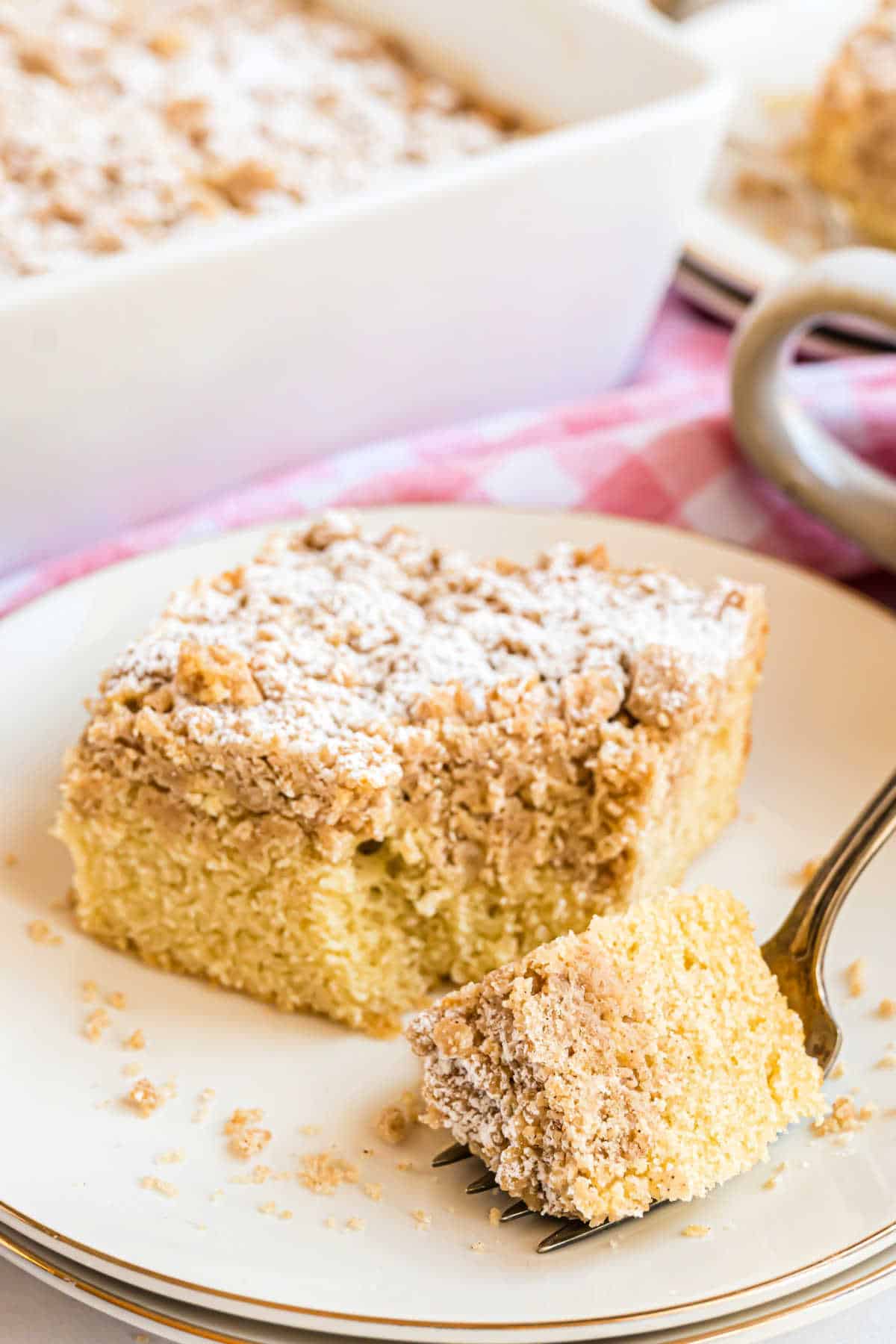 A delicious slice of crumb cake topped with powdered sugar sits on a white plate, with a fork holding a perfect bite.