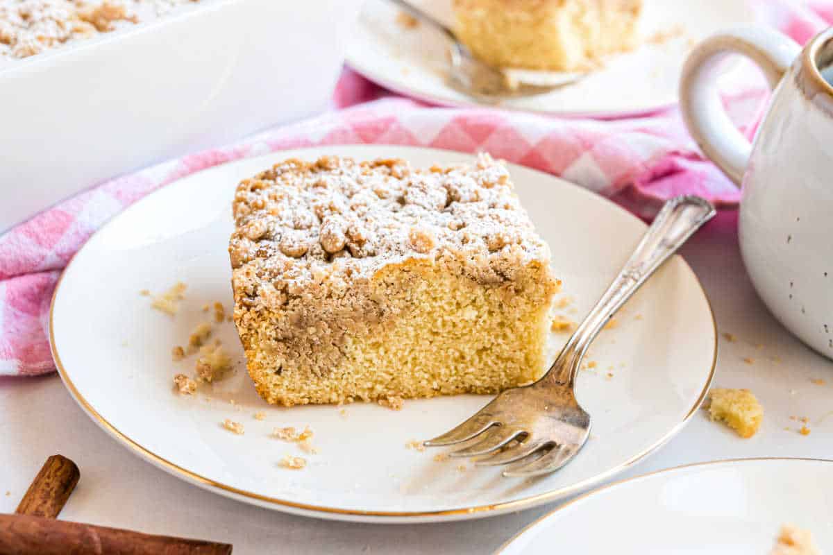 A slice of crumb cake, topped with a buttery crumb layer and dusted with powdered sugar, sits on a white plate with a fork.