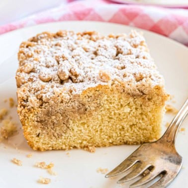 A slice of crumb cake topped with powdered sugar sits on a white plate with a fork, ready to enjoy.