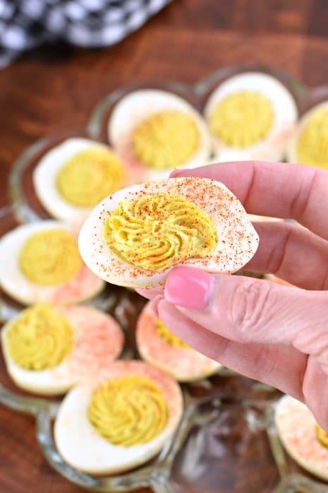 Glass plate of deviled eggs in background. Hand holding a single deviled egg.