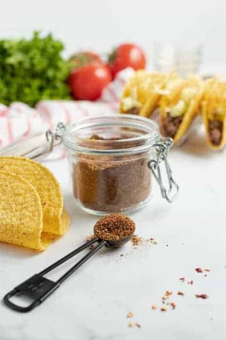 Jar of homemade taco seasoning with tacos in background.