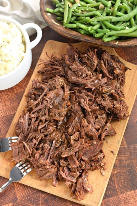 Shredded balsamic beef on wooden cutting board with green beans and mashed potatoes.