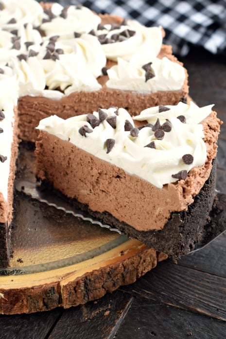 Big slice of chocolate cheesecake on pie server. Cookie crust, chocolate filling, whipped cream topping, and chocolate chips.