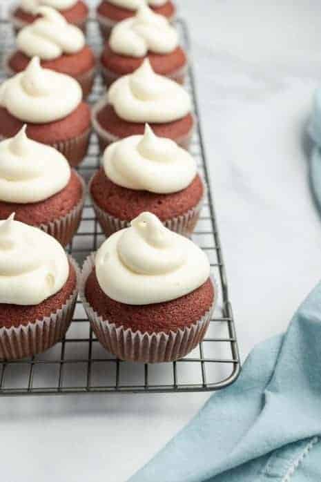 Red velvet cupcakes on a wire cooling rack topped with a swirl of cream cheese frosting.