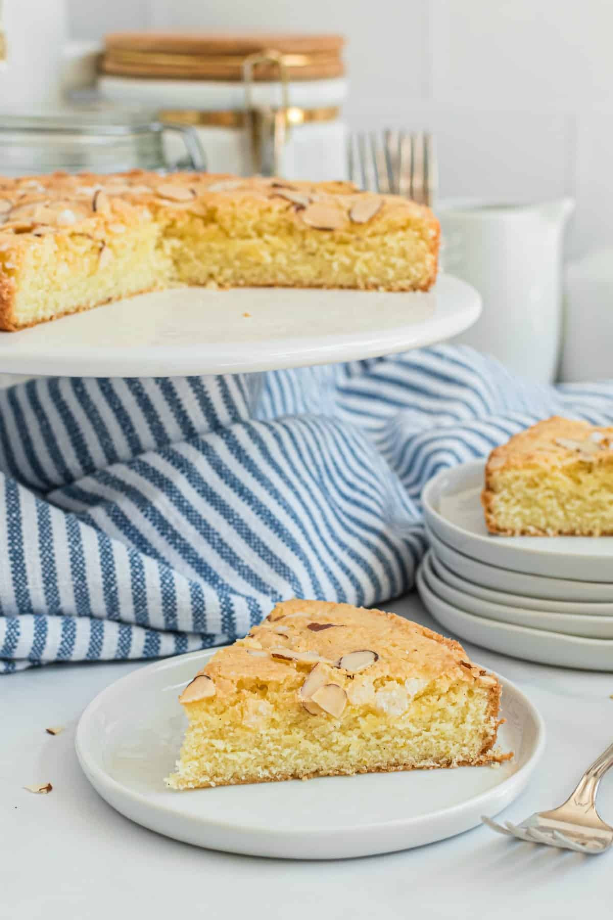 Slice of almond cake on a white plate with striped blue linen, remaining cake on a white cake platter.