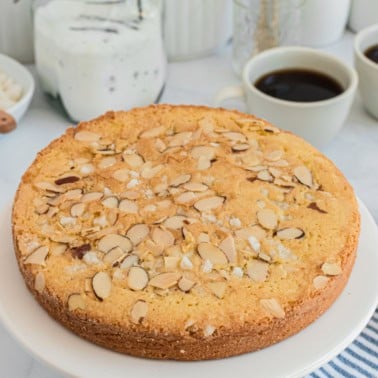 A round Swedish almond cake topped with sliced almonds sits on a white plate, with coffee cups in the background.