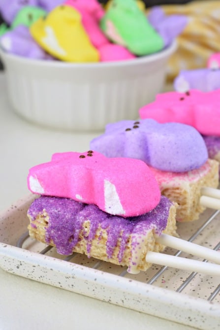 Wire rack with rice krispie treats cut into rectangles with a popsicle stick. Topped with melted white chocolate, purple and pink sprinkles, and marshmallow Peeps.