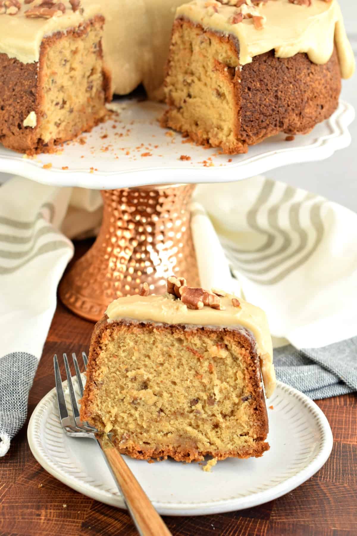 Slice of pound cake on a white plate topped with caramel glaze and pecans. Whole cake on platter in background.