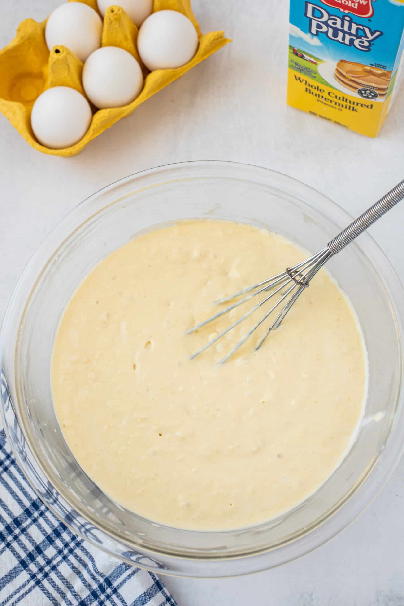 Glass bowl with whisk and pancake mix blended smooth. Eggs and buttermilk in background.