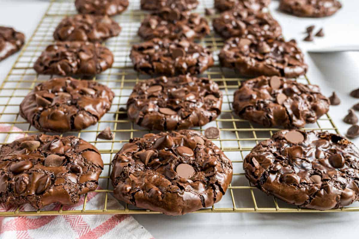 Flourless chocolate cookies with chocolate chips cooling on a wire rack, with a towel and scattered chips nearby.