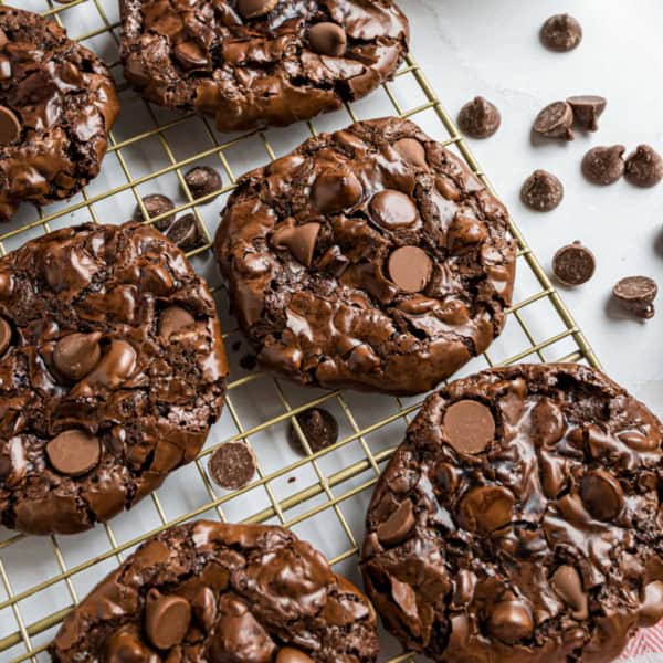 Thick, glossy flourless chocolate cookies with chocolate chips cooling on a wire rack, with chips scattered nearby.