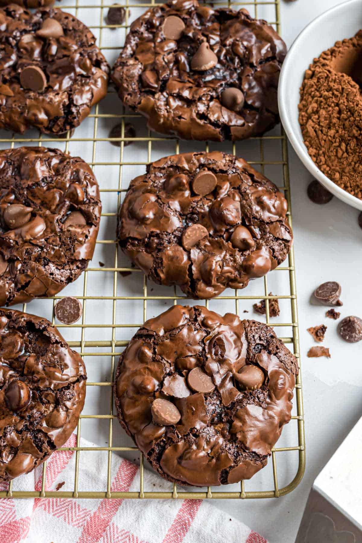 Six flourless chocolate cookies with chocolate chips on a cooling rack, next to a bowl of cocoa powder and a striped towel.