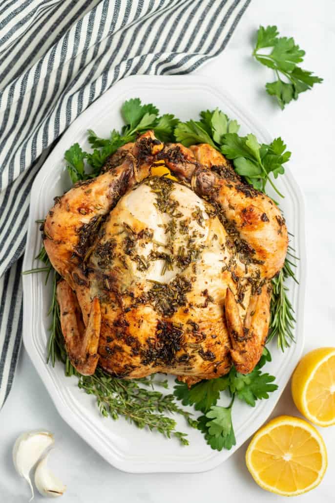 Whole roasted chicken on white plate with fresh herbs, garlic, and lemon.