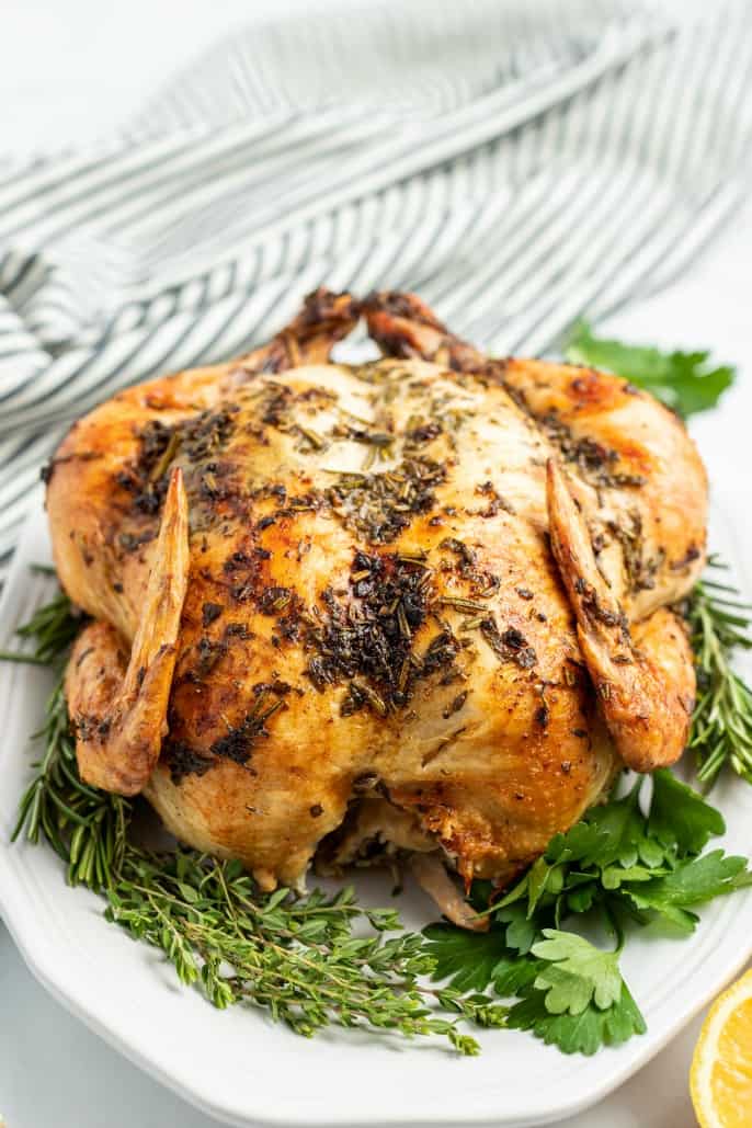 Whole chicken with garlic and herbs roasted on the skin. Served on white platter with fresh herbs.