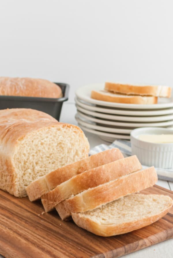 Sliced loaf of homemade white bread on bread board with a stack of plates in the background.