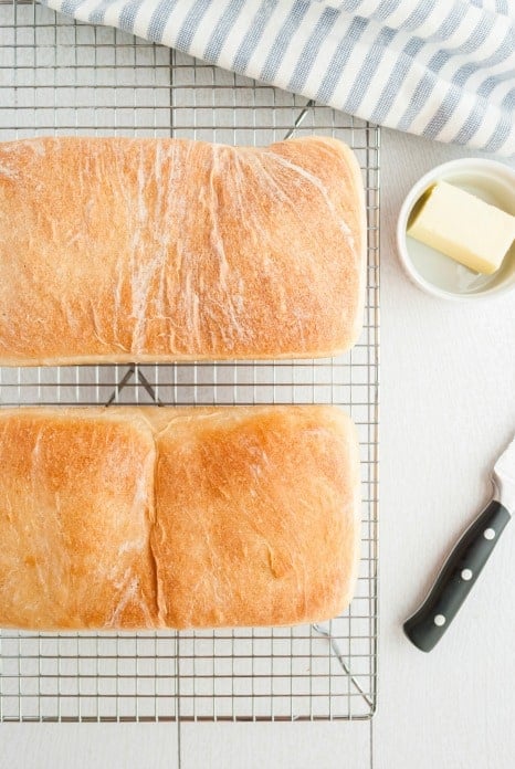 Two loaves of homemade bread on a wire rack with a bowl of butter and a knife.