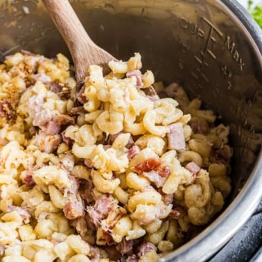 Creamy macaroni with diced ham and bacon, reminiscent of instant pot chicken cordon bleu, being stirred in a metal pot with a wooden spoon.
