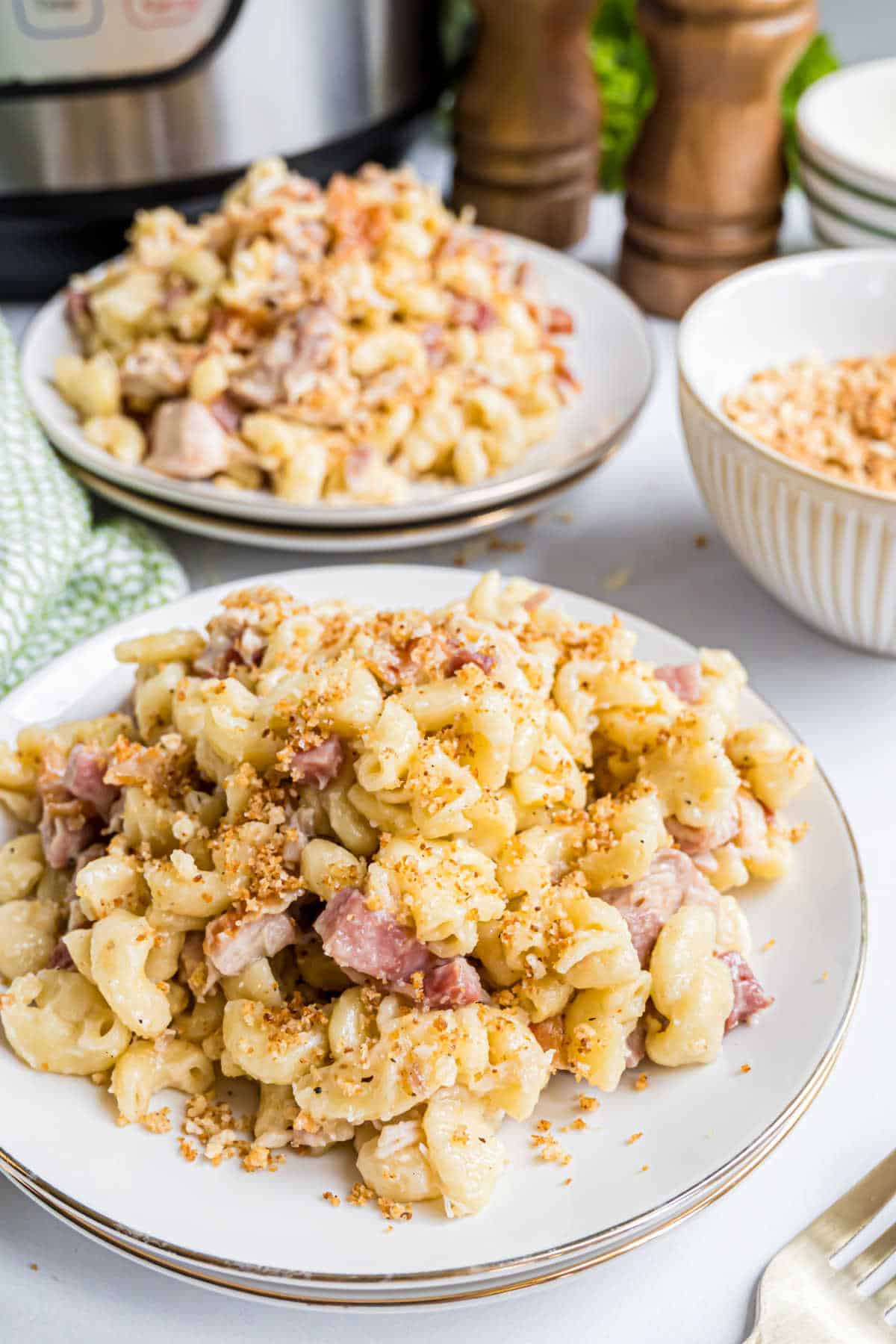 Two plates of creamy macaroni and cheese with ham and breadcrumbs, reminiscent of instant pot chicken cordon bleu, with a bowl and Instant Pot in the background.