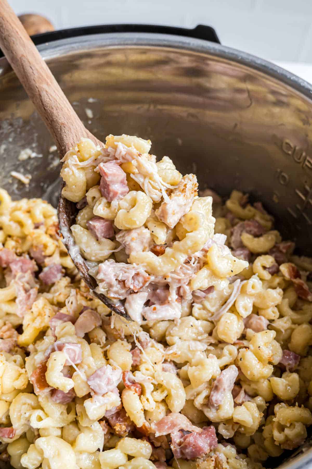 A wooden spoon scoops creamy macaroni with chunks of ham, reminiscent of instant pot chicken cordon bleu, from a pot.