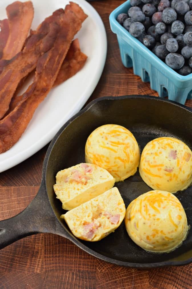 Small skillet with 4 ham and cheese egg bites, one cut in half. Side plate with cooked bacon and a small teal bowl of blueberries.