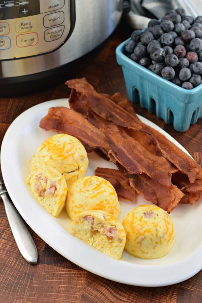 White oval plate with ham and cheese egg bites and several strips of cooked bacon. Teal berry bowl with blueberries.