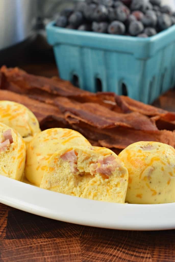 Ham and Cheese Egg bites made in the instant pot on a plate with cooked bacon and a teal bowl full of blueberries.