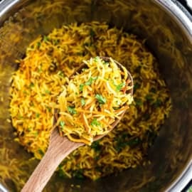 A wooden spoon holding yellow seasoned instant pot Rice-A-Roni with herbs over a pot of the same rice.