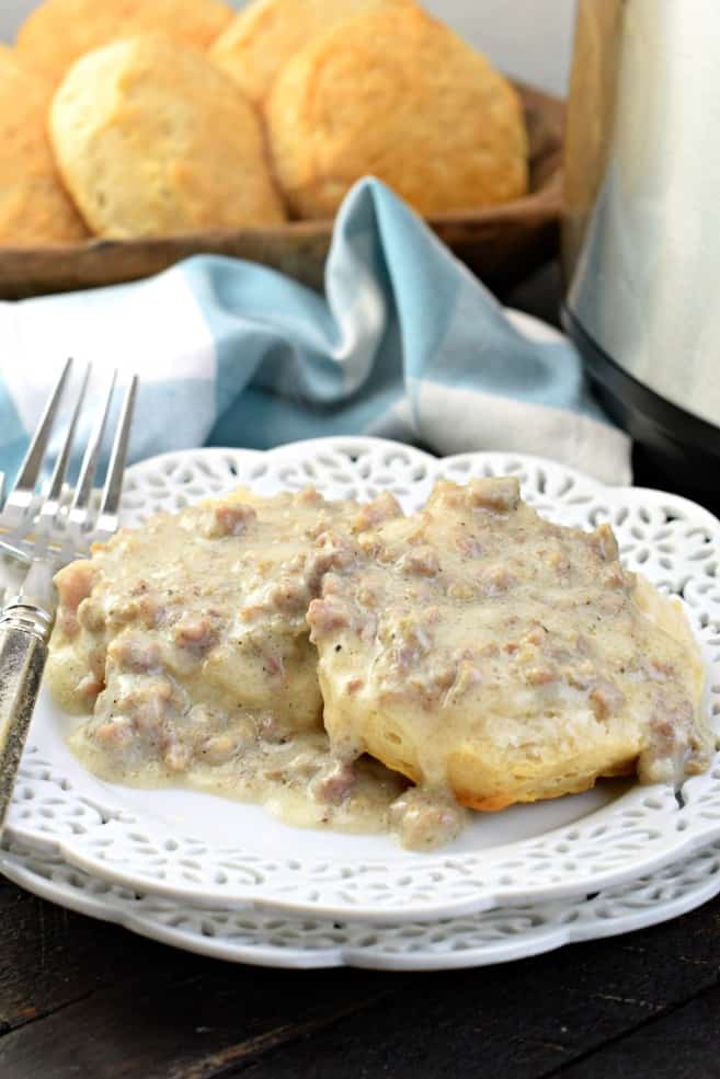 White frilled plate topped with an open faced biscuit and homemade sausage gravy. Tray of extra biscuits in background.