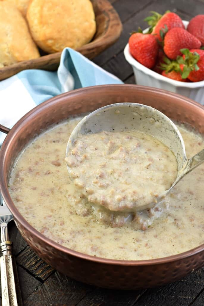 Copper bowl with sausage gravy and ladle. Bowl of strawberries and fresh biscuits in background.