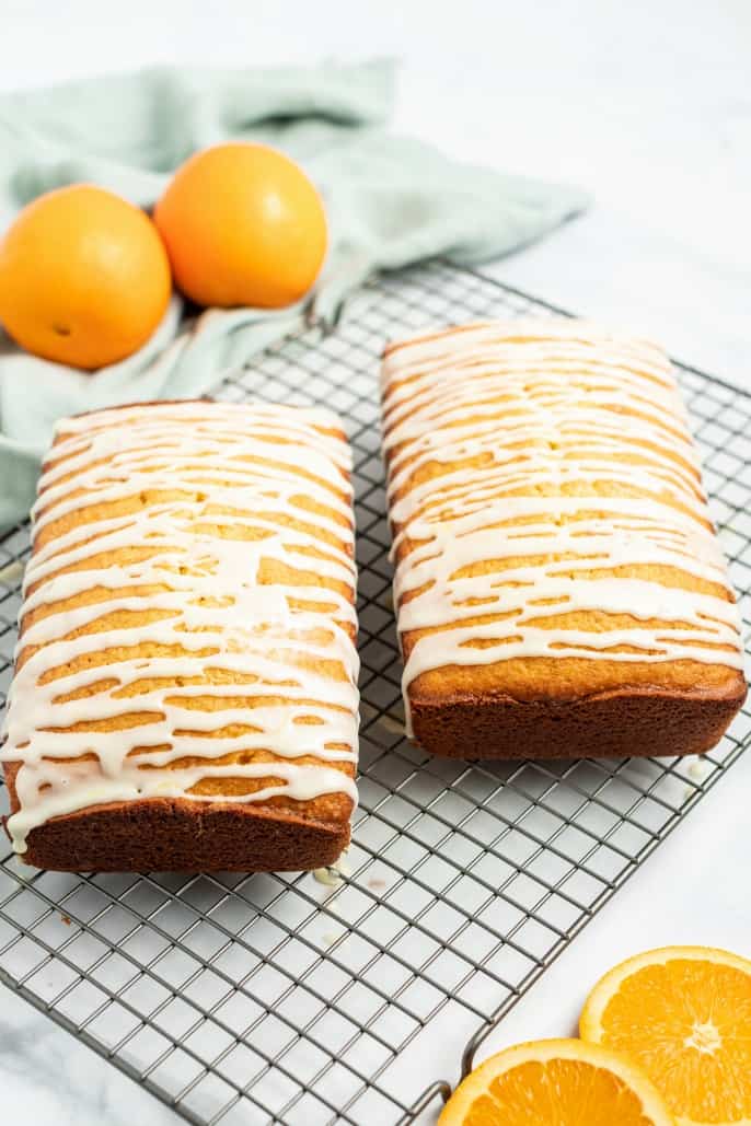 2 loaves of glazed orange bread on a wire rack with two oranges in background.