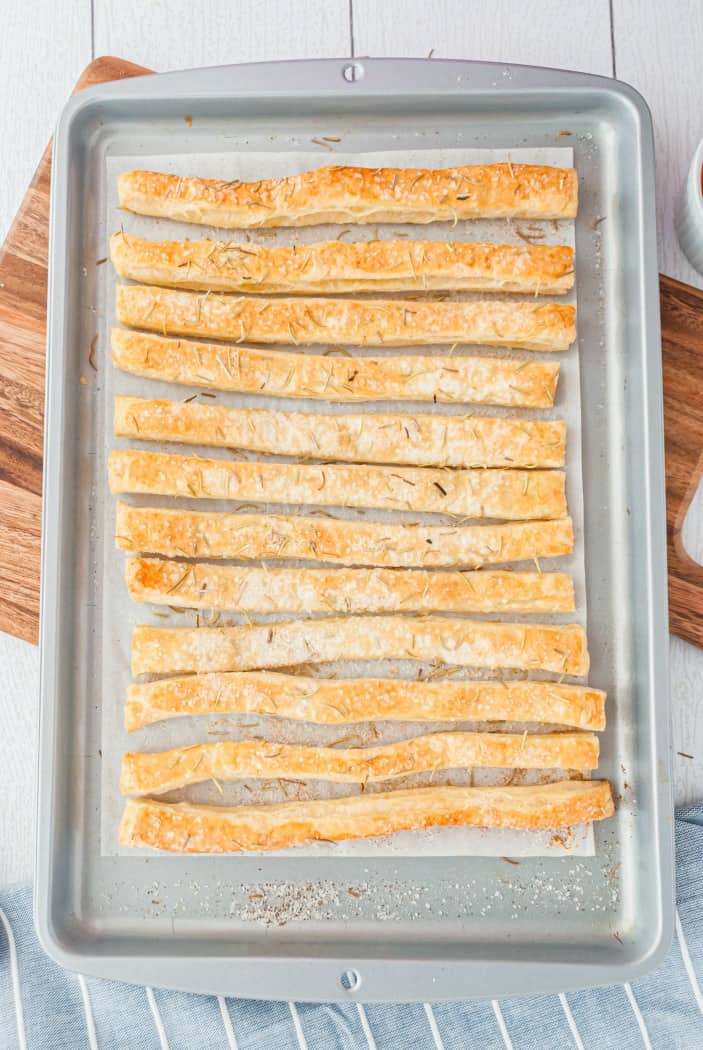 Metal baking sheet with 12 puff pastry breadsticks on parchment paper.