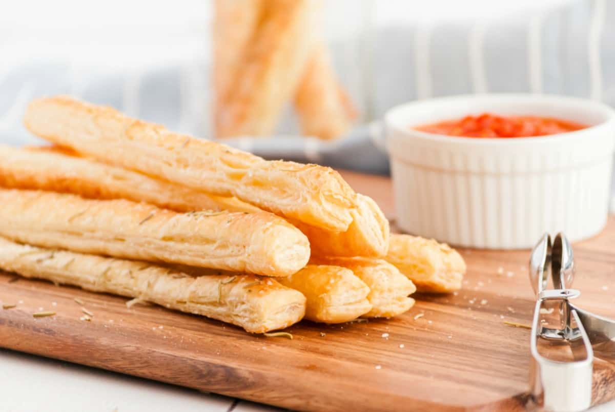 Puff pastry breadsticks on wooden board with sauce on side.