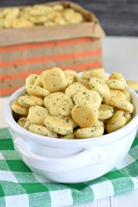 White bowl on green checkered napkin filled with dill and ranch oyster crackers. Paper bag (with orange stripes) in background filled with more snack mix.