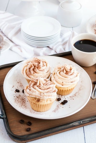 White plate with three tiramisu cupcakes dusted with espresso powder. Served on wooden tray with a cup of coffee in a white mug.