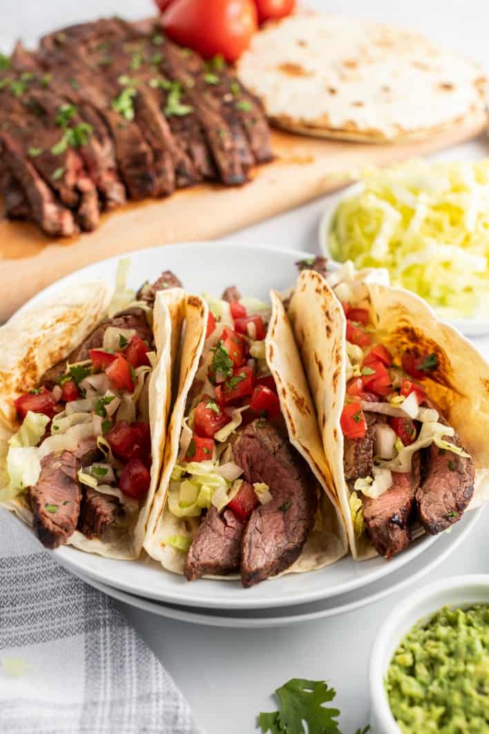 3 flour tacos topped with carne asada, pico de gallo, lettuce, and cilantro. Served on a white plate with a bowl of guacamole.