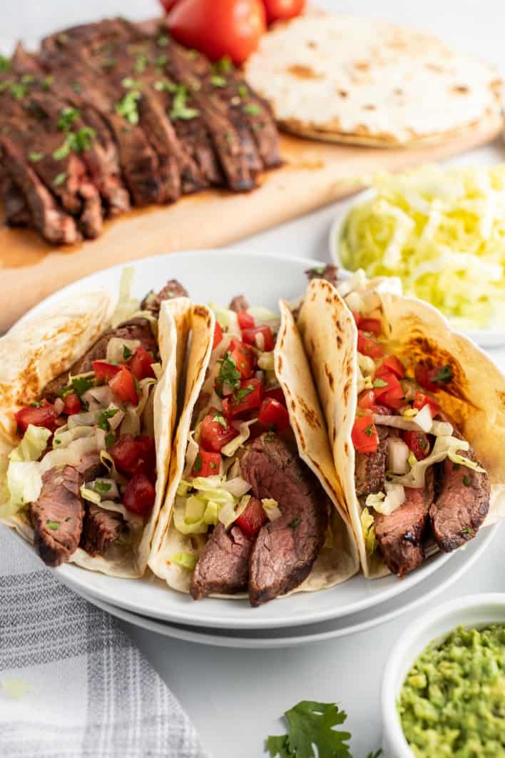 3 flour tacos topped with carne asada, pico de gallo, lettuce, and cilantro. Served on a white plate with a bowl of guacamole.