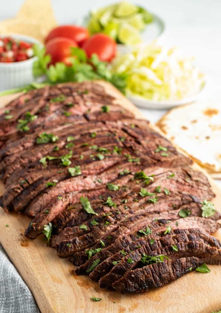 Grilled carne asada (flank steak) on a wooden cutting board with all the fixings on the side.