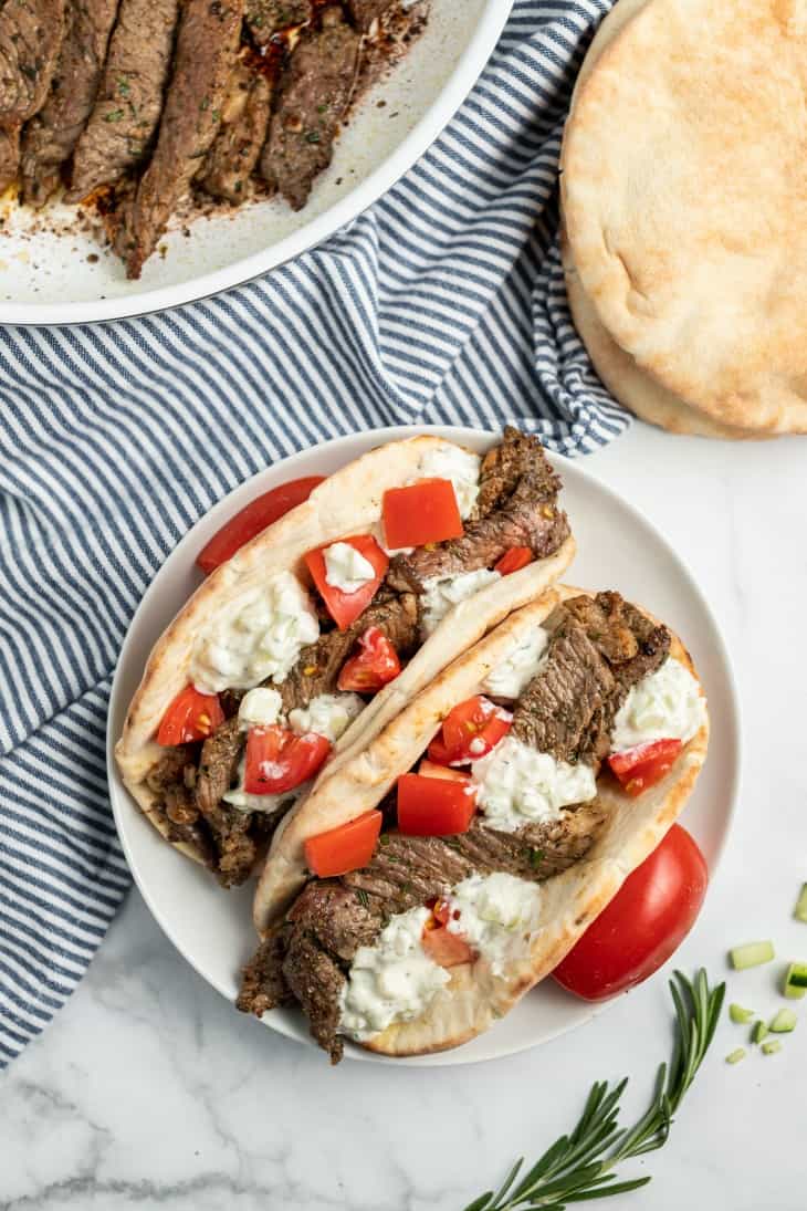 Two pitas on a white plate with greek seasoned sirloin steak, tomatoes, and cucumber sauce.