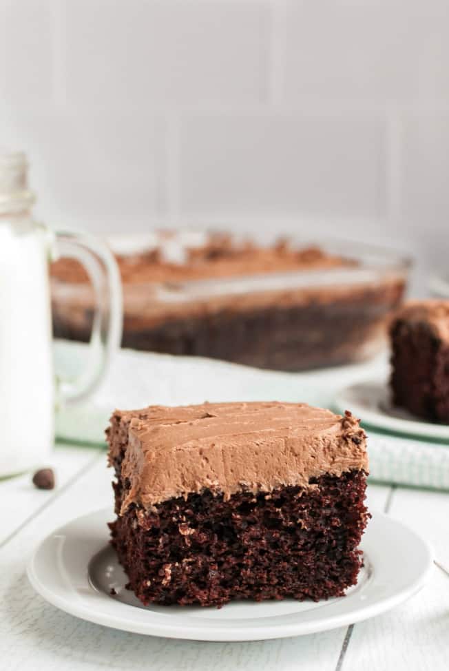 Slice of chocolate wacky cake with buttercream frosting on a white plate, with cake in background.