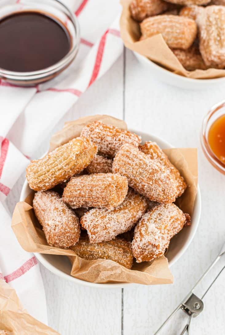 Bowl lined with parchment paper filled with churro bites. Served with a side of hot chocolate sauce and caramel.