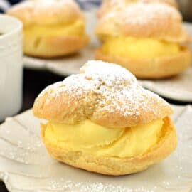 Cream puff on a white plate filled with custard and topped with powdered sugar.