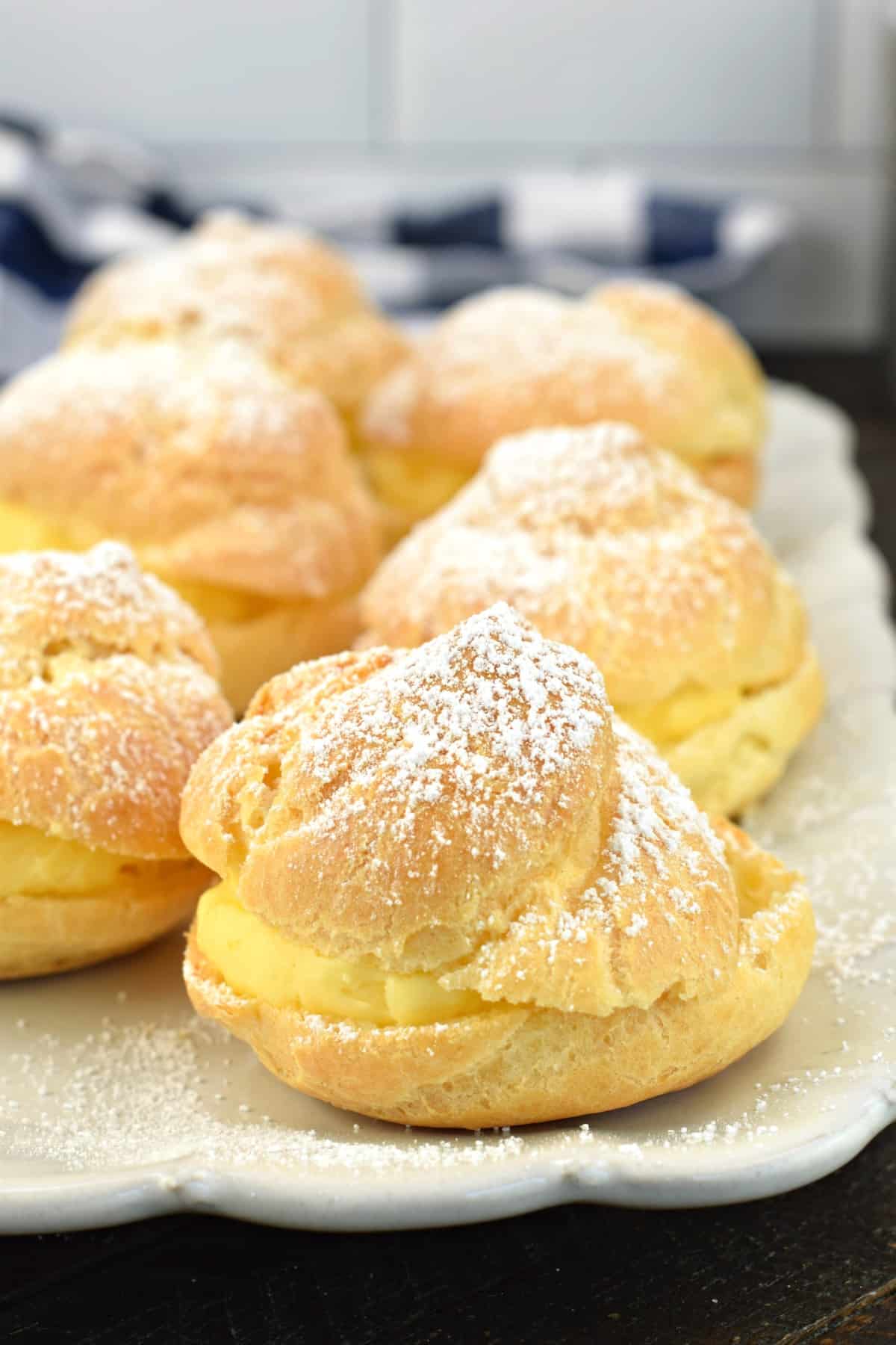 6 cream puffs on a white serving plate topped with powdered sugar.
