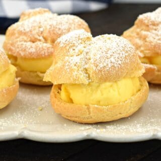 Cream puffs with vanilla custard filling and topped with powdered sugar on a white serving plate.