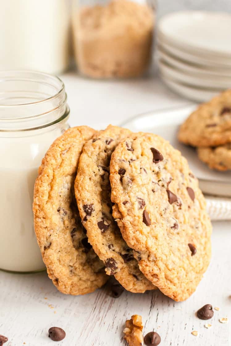 Three large chocolate chip cookies stacked vertically against a clear mug of milk.