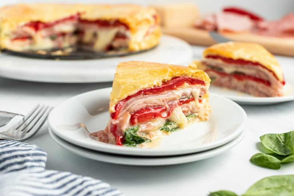 Slice of Italian sandwich torte on a stack of white plates, with cheese oozing out.