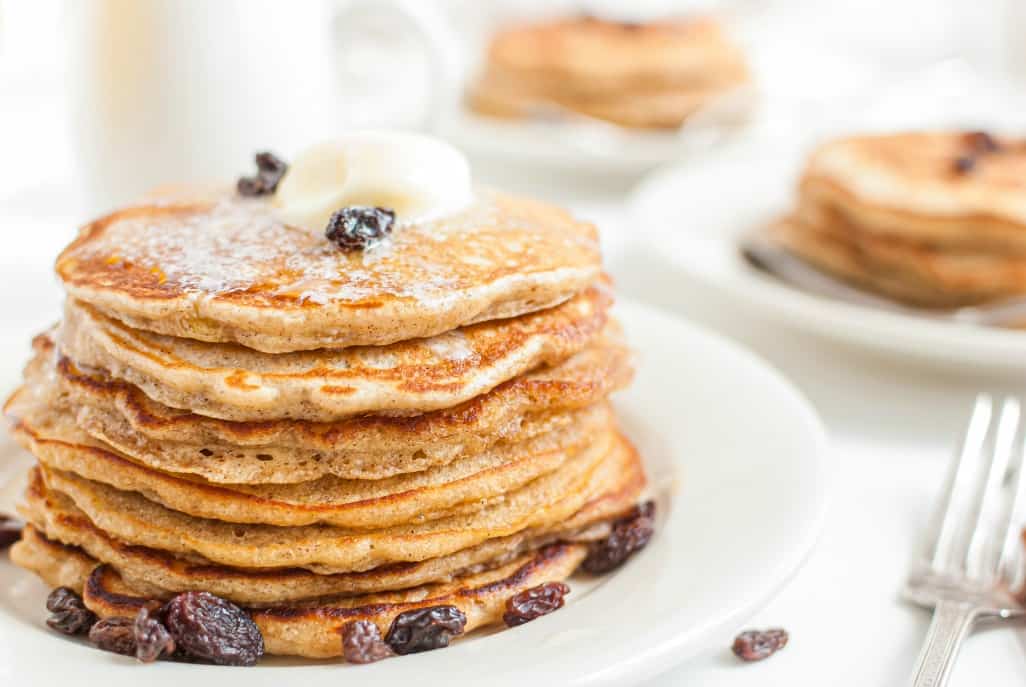 Stack of oatmeal cookie pancakes with butter, cinnamon, and raisins on a white plate.