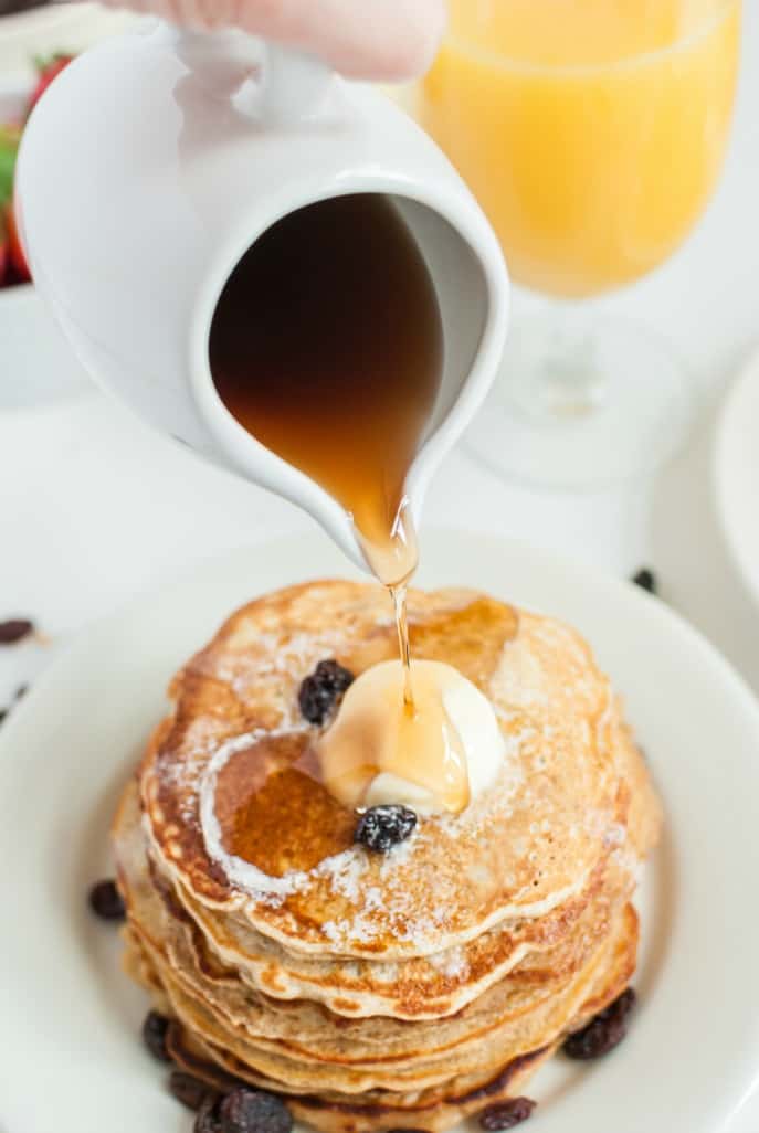 Stack of pancakes with butter and raisins and syrup being poured over the top.