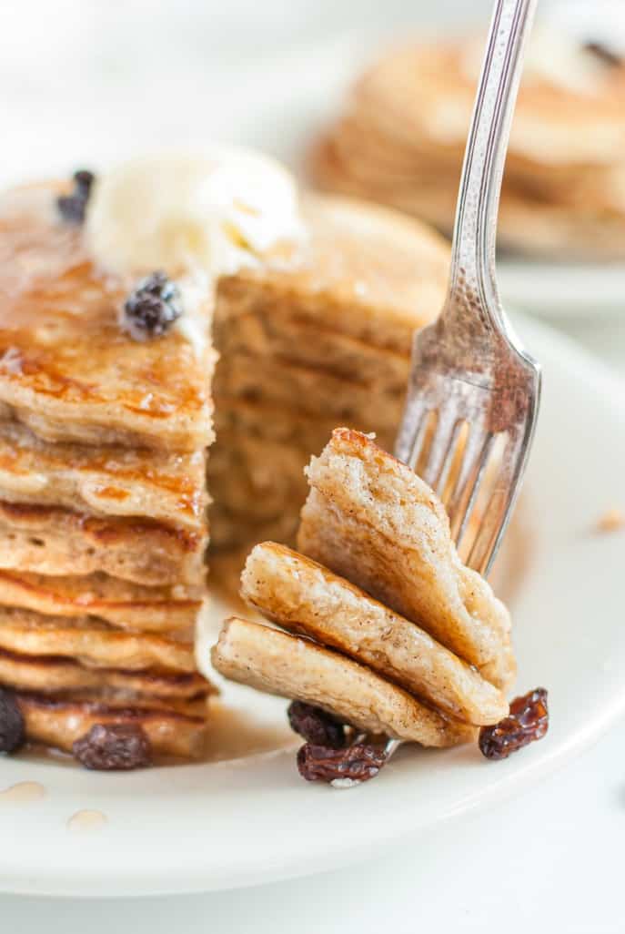 Stack of pancakes with one bite cut and being lifted by a fork. Raisins on the side.