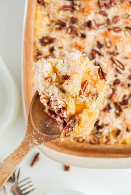 Rectangle baking dish with peach dump cake featuring wooden spoon lifting cake out of pan. Topped with coconut and pecans.